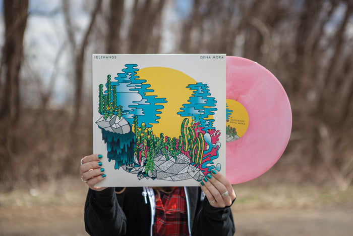 A person with teal-painted nails holds an IDLEHANDS Dena Mora vinyl. The colorful cover features abstract plants, rocks, and a bright yellow circle. This limited edition LP is displayed outdoors against a backdrop of blurred trees.