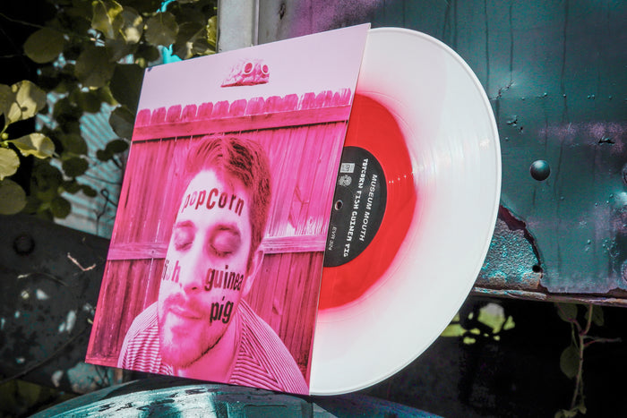 A Limited Pressing red and white vinyl by Museum Mouth is partly covered by an album cover with a man's face and text like "popcorn" and "Guinea Pig LP," set outdoors among plants on a green metal surface.
