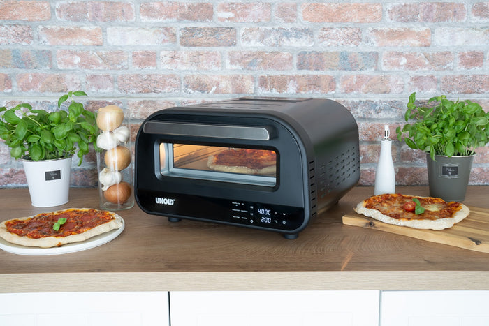 Der Unold B-Ware Pizzaofen Don Luigi in Schwarz steht auf einer Holzfläche neben zwei Basilikumpflanzen, Zwiebeln, Salz- und Pfefferstreuern und zwei fertigen Pizzen auf Schneidebrettern, im Hintergrund eine Backsteinmauer.