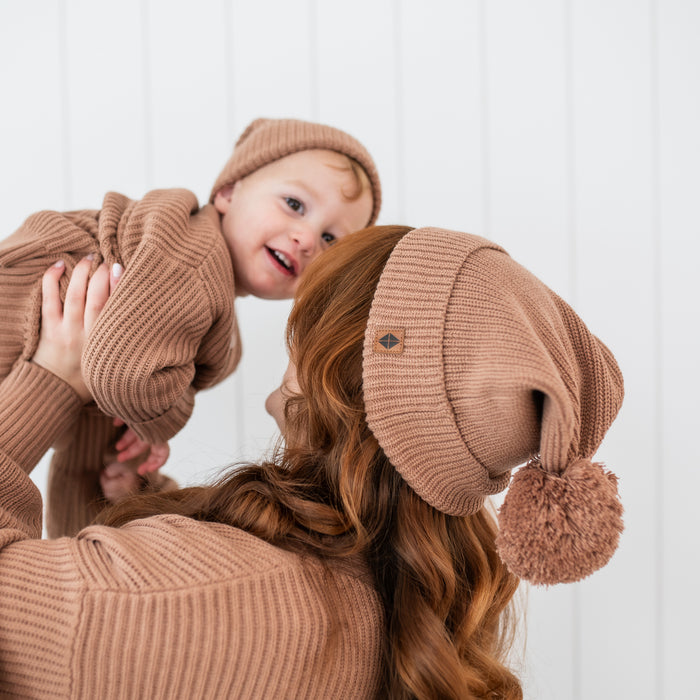 Mother holding her son while wearing the Chunky Knit Adult Pom Beanie in Latte