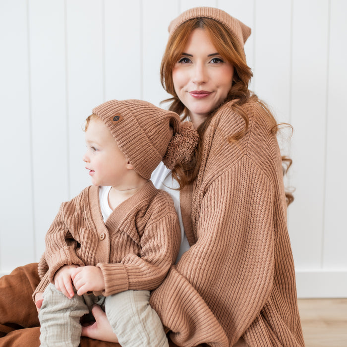 Young toddler wearing the Chunky Knit Pom Beanie in Latte and matching chunky knit oversized cardigan sitting on his mom's lap who is wearing matching colored chunky knit items