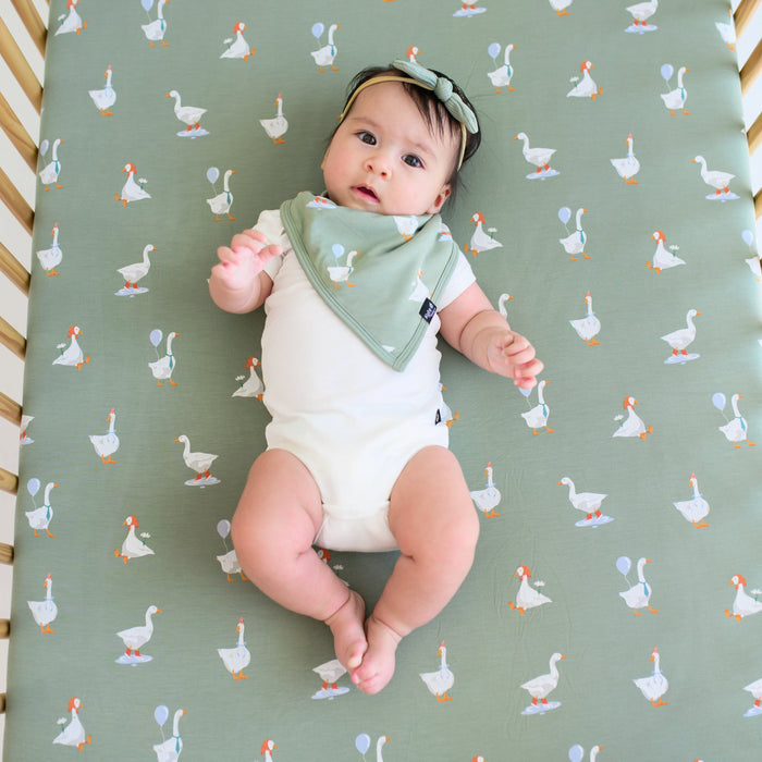 Infant laying in a crib on a Crib Sheet in Silly Goose wearing a matching bib, Cloud Bodysuit and ribbed tiny nylon bow in Thyme