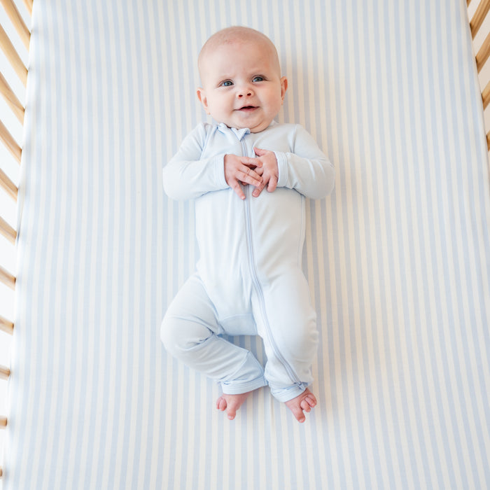 Smiling infant laying in a crib on top of a Crib Sheet in Small Mist Stripe wearing a Mist Zippered Romper