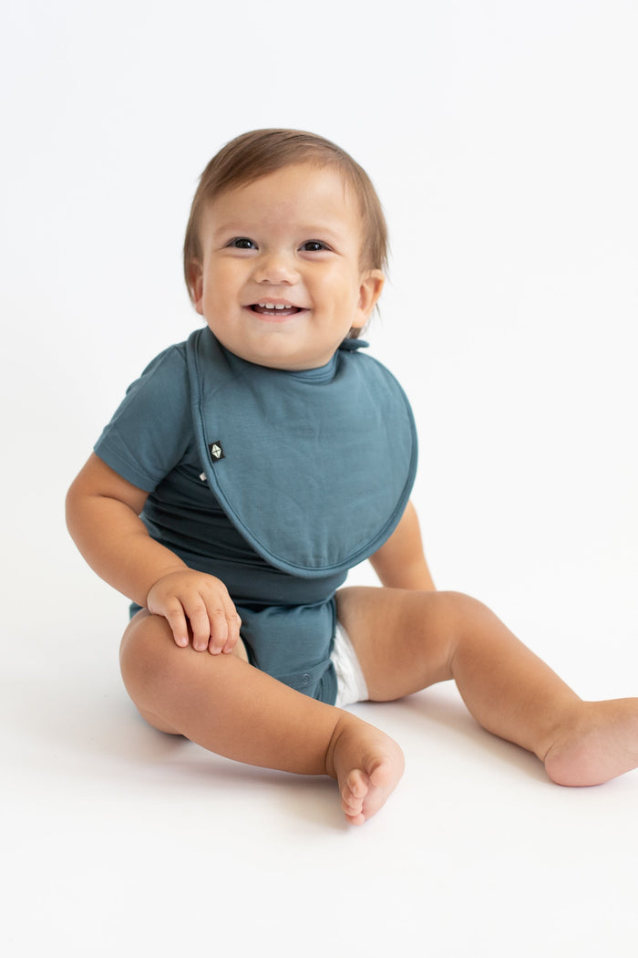 Smiling infant sitting down wearing the Cushy Bib in Atlantic with matching short sleeve bodysuit