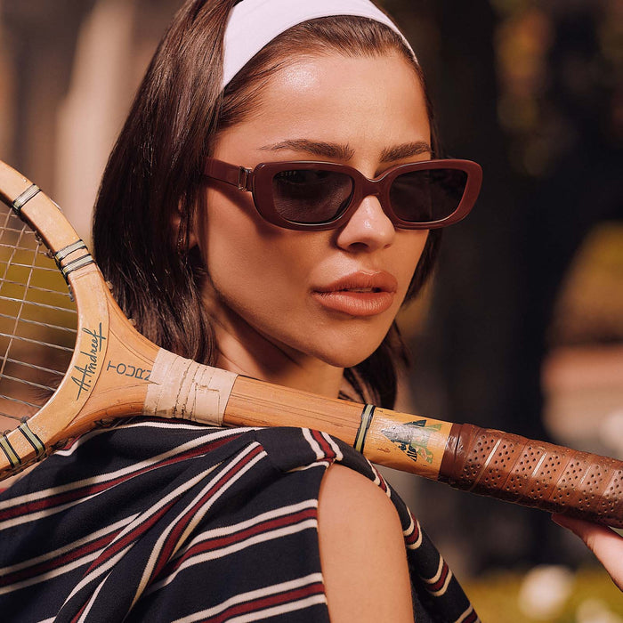 lifestyle - female with a tennis racket wearing diff eyewear featuring the daphne rectangle sunglasses with a rugby red acetate frame and grey polarized lenses angled view