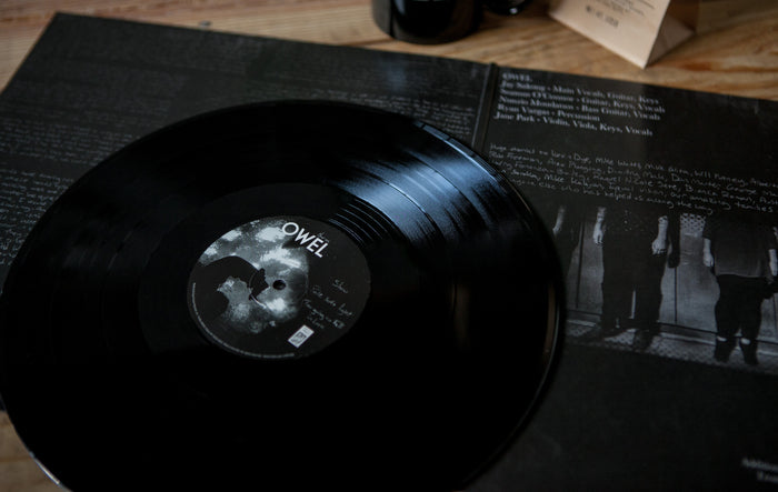 A limited edition black double LP vinyl of OWELs Dear Me rests on its open cover, showcasing lyrics and a black-and-white band photo, all placed on a wooden surface.