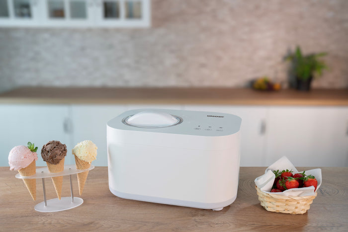 Die Unold Eismaschine Snow 1 L in Weiß steht auf einer Küchentheke neben einem Ständer mit drei Eistüten und einem Korb mit frischen Erdbeeren. Im Hintergrund ist eine verschwommene Küche zu sehen.
