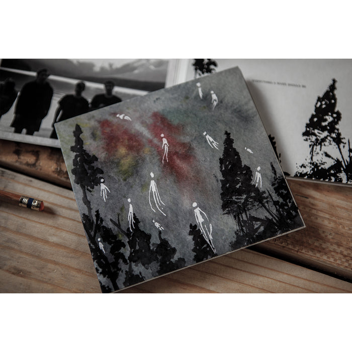 A Household postcard featuring painted black trees and ghostly white figures rests on a wooden surface beside a pencil, open black-and-white books, and what looks like the track list for the music album "Dark Matter.