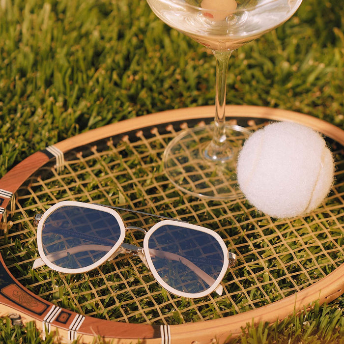 still life of diff eyewear featuring the joey round sunglasses with a courtside ivory & gold metal frame with blue slate gradient flash lenses on top of a tennis racket