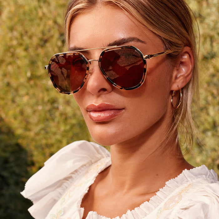 lifestyle - blonde female in her backyard wearing diff eyewear featuring the joey round sunglasses with a espresso tort & gold metal frame with brown gradient polarized lenses front view