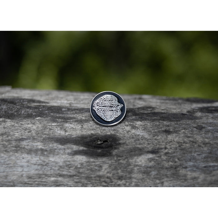 A Gatherers black lapel pin featuring a striking white geometric pattern rests on a weathered wooden surface, its bold lines distinct against the blurred green background.