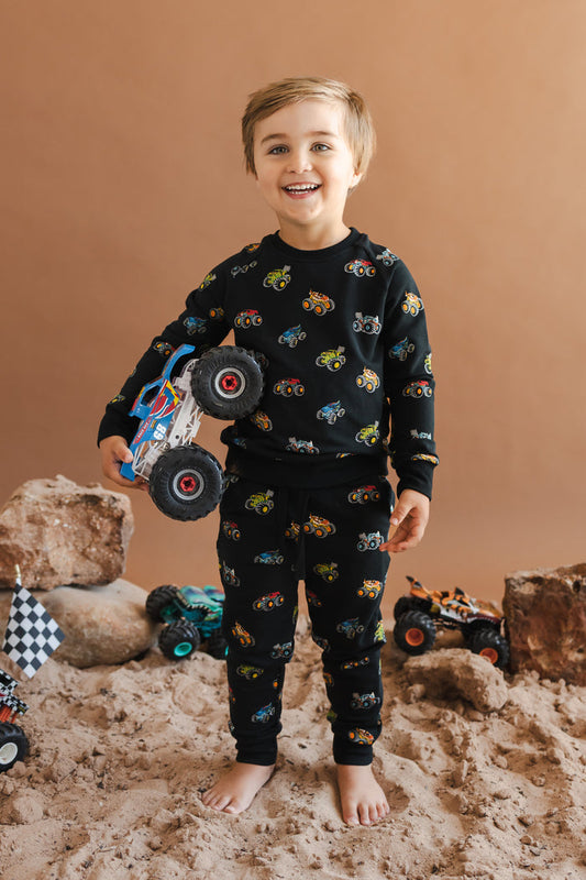 Toddler wearing Black Long Sleeve Jogger Set in Monster Truck print holding a truck standing on sand and rock setup