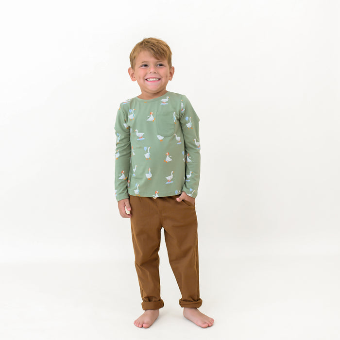 Smiling boy looking off to the side standing with one hand in the pocket of his brown pants wearing the Long Sleeve Toddler Crew Neck Tee in Silly Goose
