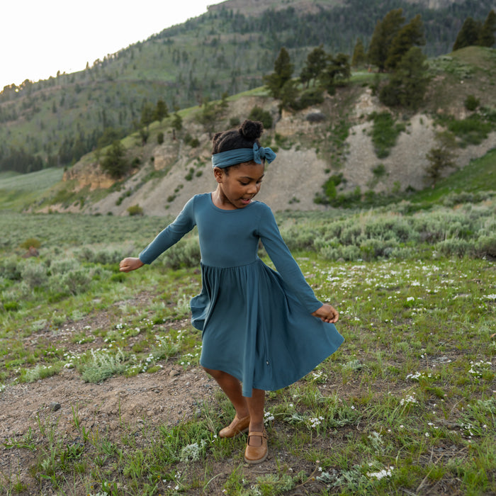 Girl twirling in a field wearing the Long Sleeve Twirl Dress in Atlantic paired with a matching bow headband