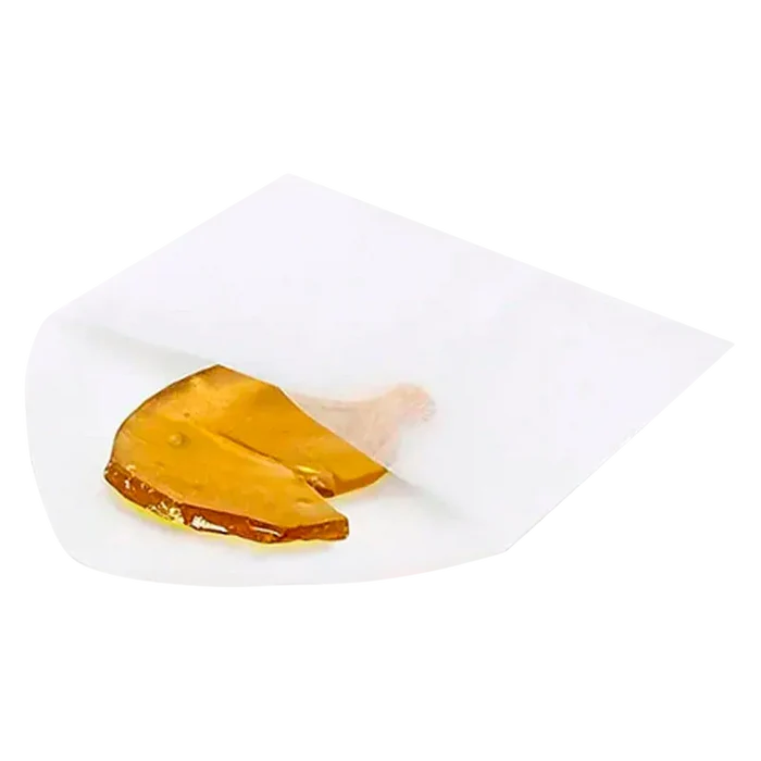 Angled close-up of amber shatter partially wrapped in white parchment paper, labeled Max Exhaust Haze Shatter.
