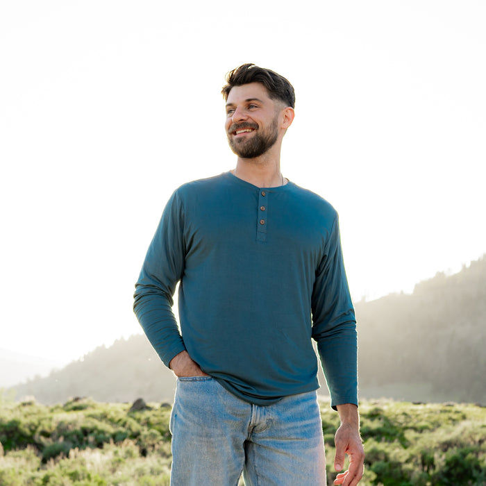 Male model standing in a field wearing the Men's Long Sleeve Henley Top in Atlantic with one hand in his jean pocket