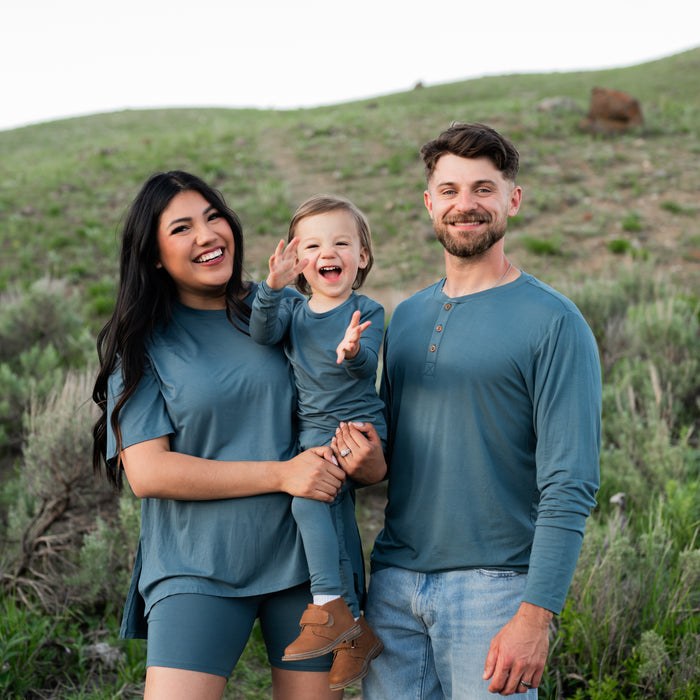 Family matching in Atlantic standing in a field. Pictured is the Men's Long Sleeve Henley Top in Atlantic, Toddler Long sleeve pajama set and Women's biker short set