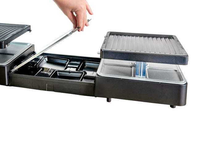 Eine Hand hebt die obere Grillplatte des Unold Raclette Extendable an, einem schwarzen Elektrogrill mit mehreren Fächern und Ablagen, abgebildet vor einem weißen Hintergrund.