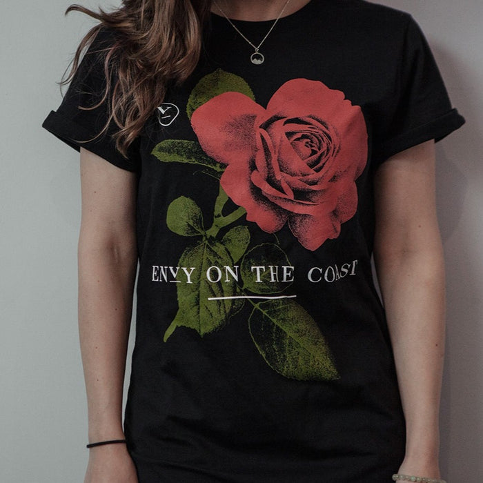 A person with long brown hair wears Envy On The Coast’s Black Rose T-Shirt, showcasing a bold red rose graphic, green leaves, and the band name on the front. They finish their look with a stylish necklace.