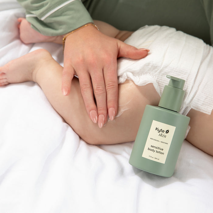 Person holding a child's leg with a bottle of Kyte Skin sensitive body lotion on a white surface