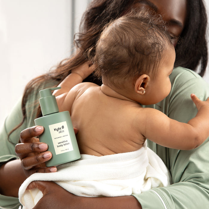 Mom holding baby wrapped in a towel and holding a bottle of Kyte Skin sensitive body lotion