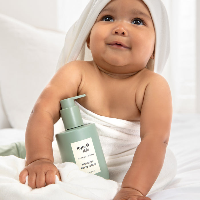 Baby holding a bottle of Kyte Skin sensitive body lotion with a white towel on head.