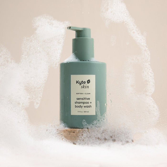 Green bottle of Kyte Skin soap and body wash with bubbles on a beige background