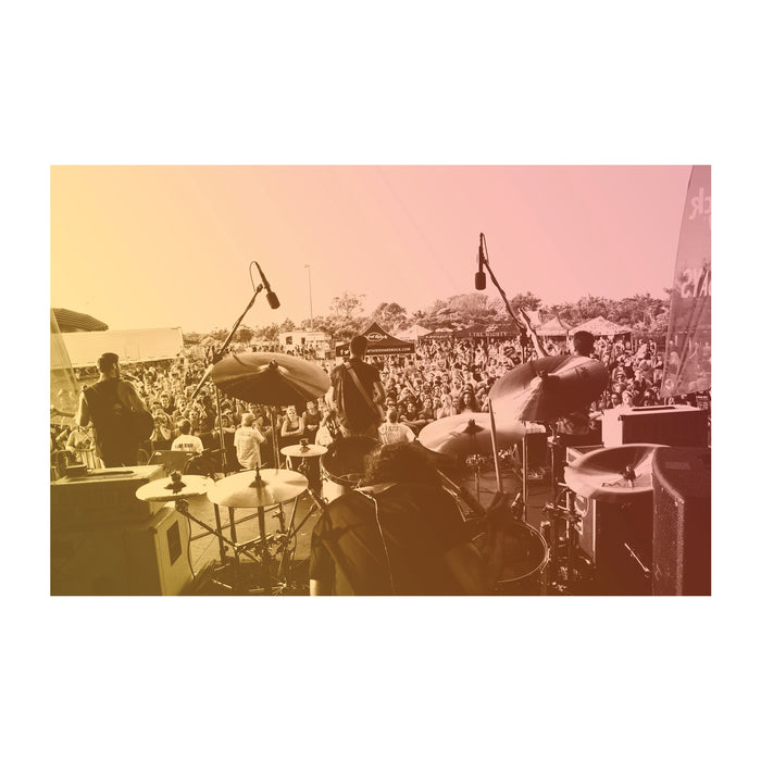 The band I The Mighty performs on an outdoor stage before a large daytime crowd, with drums and microphones in the foreground, all seen through a warm gradient color overlay.