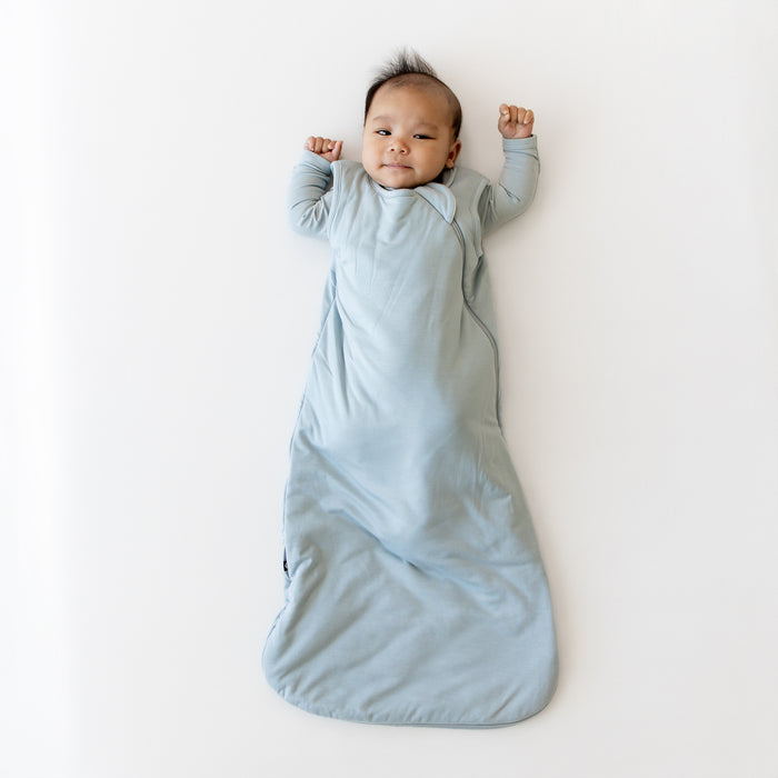 Baby in fog blue sleep bag and matching footie on white surface