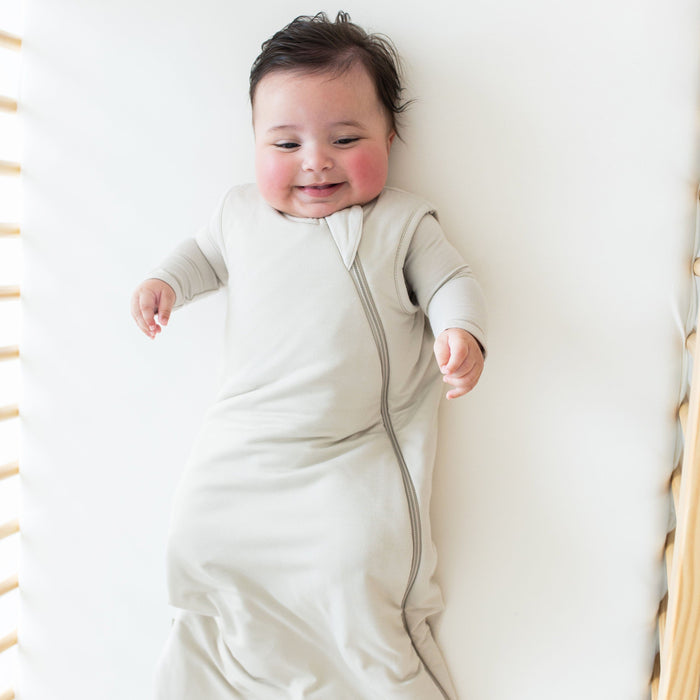 Smiling baby laying in a crib wearing Kyte Baby Sleep Bag in Oat 2.5