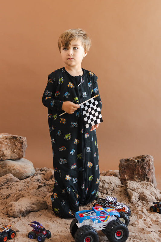 Boy wearing black Sleep Bag Walker with Monster Truck print standing on sand and rocks