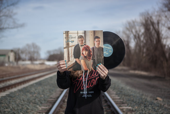 A person stands on railroad tracks holding a Merriment Sway LP Black Limited Pressing in front of their face, obscuring their identity. Their blue-painted nails clutch the record, which displays three people posing on the cover.