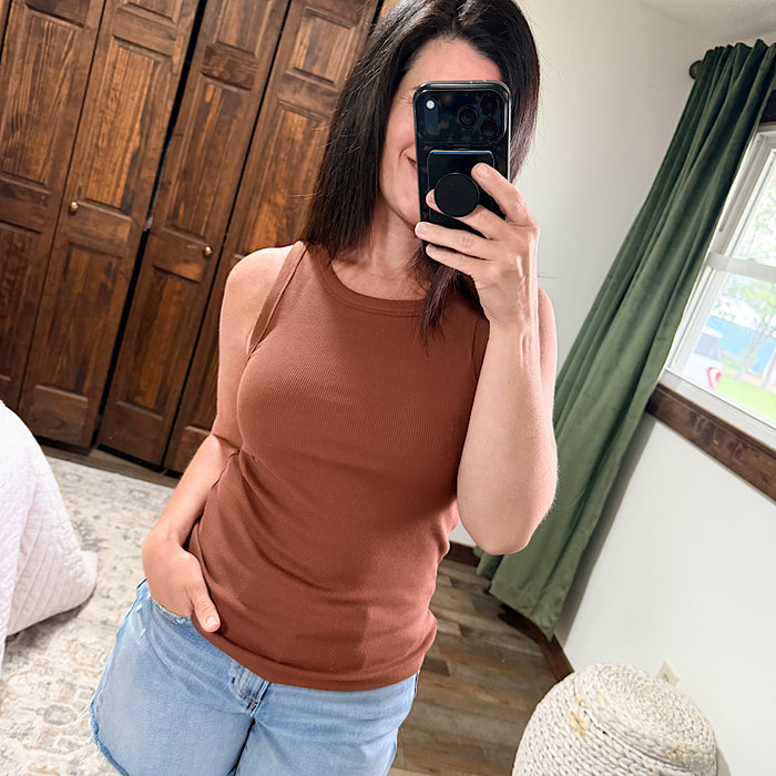 Woman taking a mirror selfie wearing a brown tank top and light blue jeans in a room with wooden cabinets and green curtains.