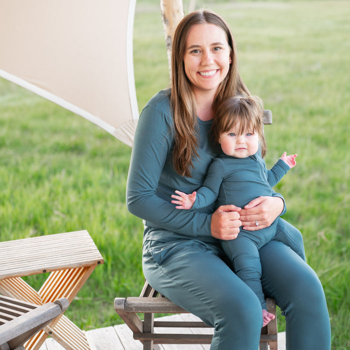 Female model sitting on a wooden stool wearing the Women's Jogger Pajama Set in Atlantic holding infant child wearing a matching zippered romper