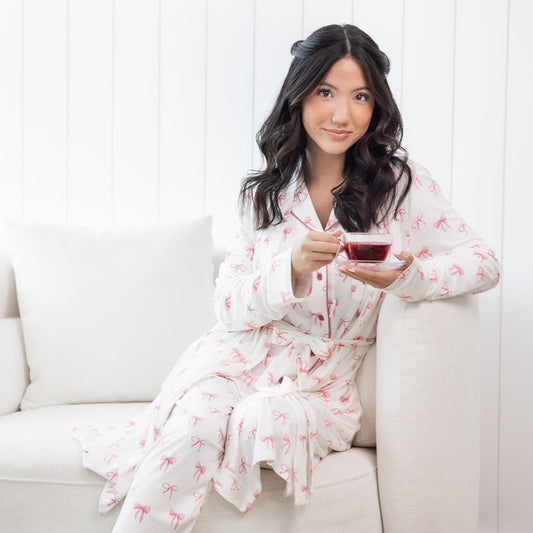 Woman wearing white long sleeve button up pajamas and robe with a pink bow print  holding a cup of tea