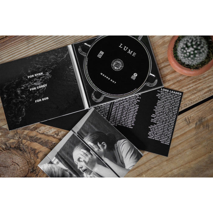 An open CD case on a wooden table displays the black Lume CD, a booklet with dedications, track list, song lyrics, and a black-and-white photo of a woman. A small potted cactus sits in the top right corner.
