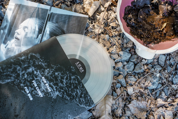 A limited pressing clear vinyl LP by Lume, titled Wrung Out, partially slides from its black and white wave-printed sleeve on gravel and dry leaves, with a monochrome photo print and a pink bowl holding dark dried leaves nearby.