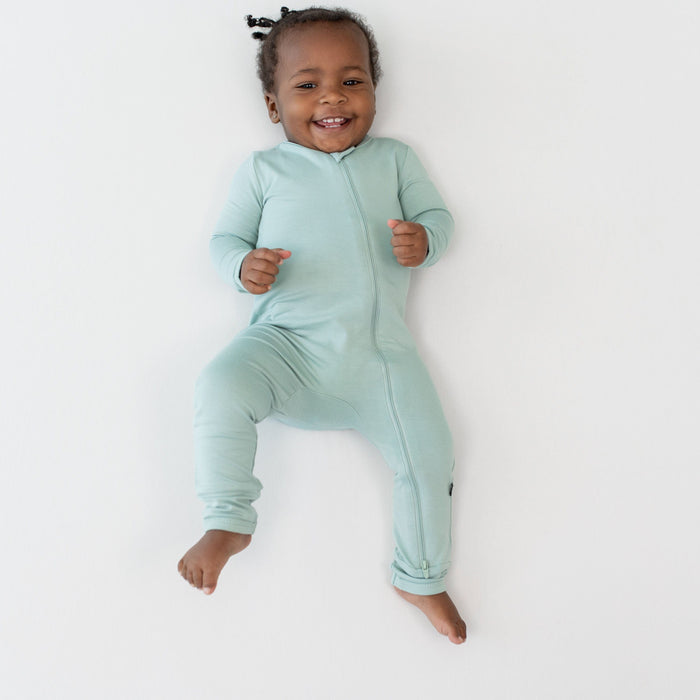 Smiling baby wearing a Zippered Romper in Sage while laying on a white surface